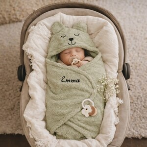 Pode incluir: Um beb&eacute; a dormir enrolado numa toalha com capuz macia e verde com tema de urso. A toalha &eacute; bordada com o nome "Emma". Uma chupeta est&aacute; na boca do beb&eacute; e um mordedor de madeira est&aacute; preso &agrave; toalha. O beb&eacute; est&aacute; numa cadeira auto cinzenta.