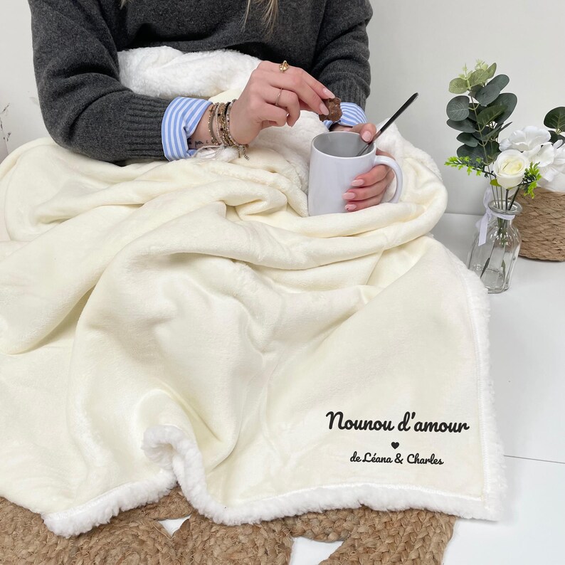 Może przedstawiać: Kremowy pluszowy koc z białym wykończeniem z sherpy. Na kocu znajduje się napis "Nounou d'amour" oraz imiona "de L&eacute;ana & Charles" wydrukowane na czarno. Osoba trzyma kubek i kawałek czekolady.