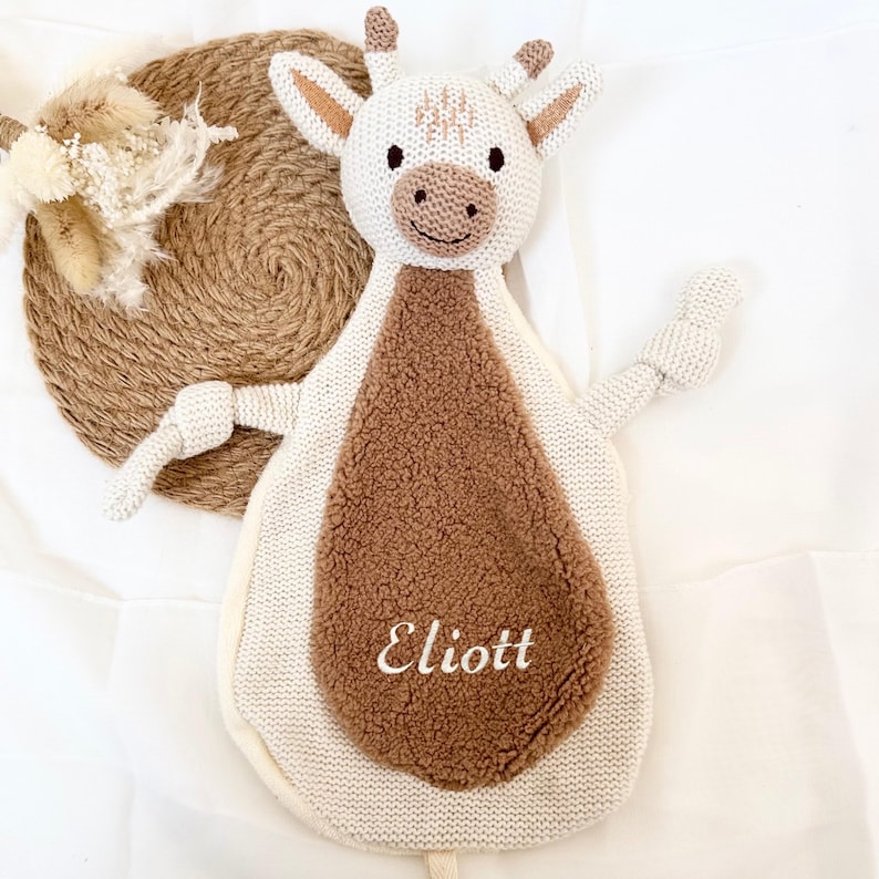 May include: White knitted plush toy shaped like a cow with brown fur on the back. The toy has a smiling face with black eyes and a black nose. The toy has a brown embroidered name "Eliott" on the back.