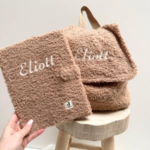 May include: A brown fuzzy backpack with a zippered pocket and the name "Elliott" embroidered on the front. The backpack is sitting on a wooden stool.
