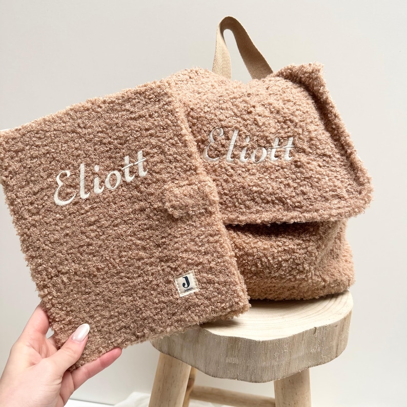 May include: A brown fuzzy backpack with a zippered pocket and the name "Elliott" embroidered on the front. The backpack is sitting on a wooden stool.