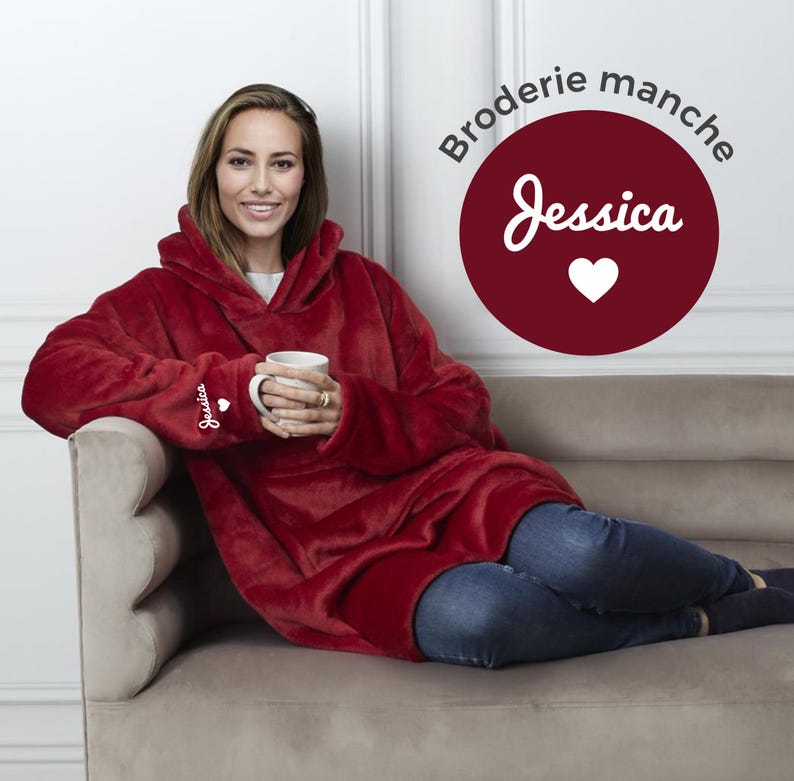 May include: A red hooded blanket with the word "Jessica" embroidered on the sleeve. The blanket is worn by a person sitting on a couch.