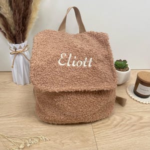 May include: A light brown fuzzy backpack with the name "Eliott" embroidered on the front.