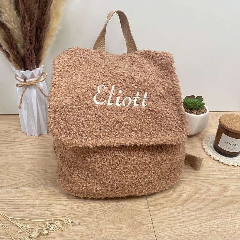 May include: A light brown fuzzy backpack with the name "Eliott" embroidered on the front.