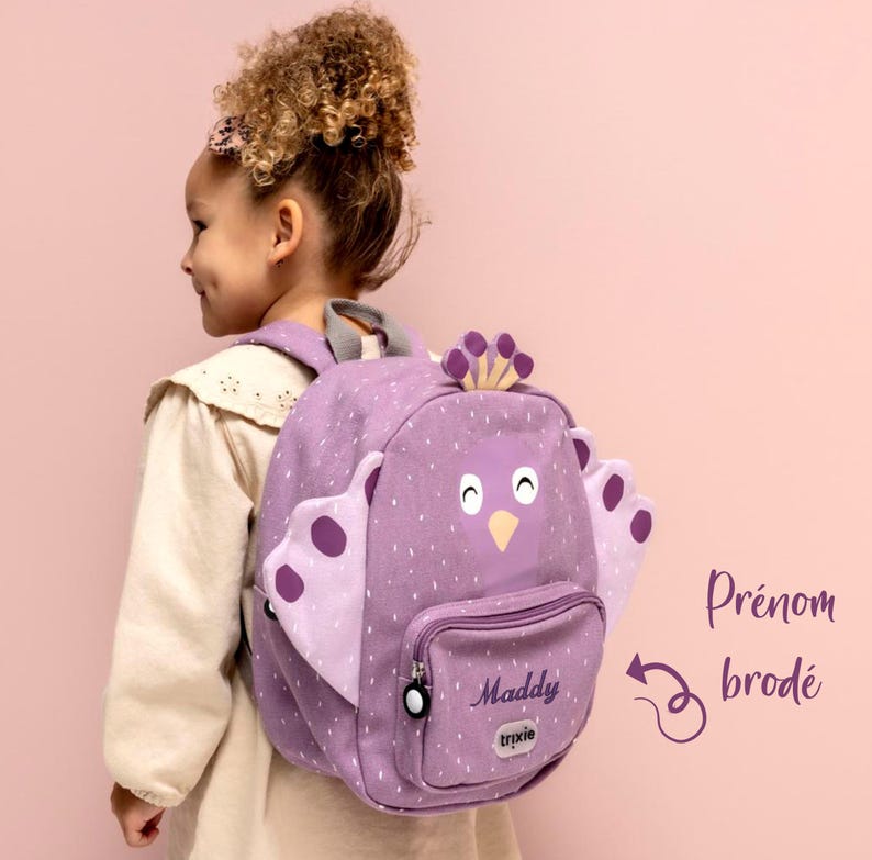 May include: A lavender backpack with a bird design, featuring a front pocket and a top handle. The backpack has white polka dots and the name "Maddy" embroidered on the front. The backpack has a bird face with a yellow beak and a crown.