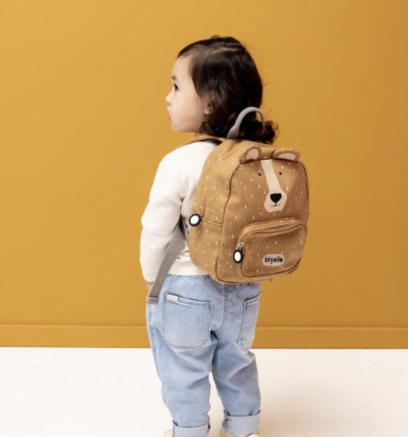 May include: A tan and white polka-dot backpack with a bear face design. The backpack has a front pocket, bear ears, and the brand name "trixie" printed on the front. The backpack is being worn by a child.