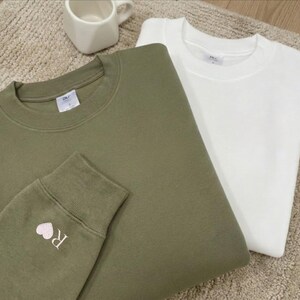 May include: Two sweatshirts, one in olive green and the other in white, are folded. The olive green sweatshirt features a small embroidered heart and the letter "R" on the cuff. A white ceramic mug is visible.