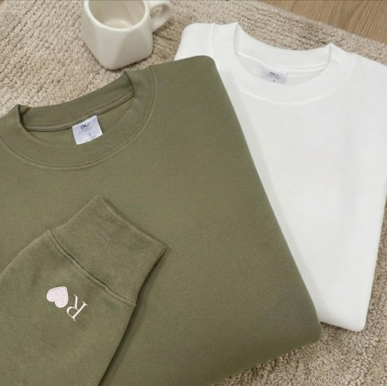 May include: Two sweatshirts, one in olive green and the other in white, are folded. The olive green sweatshirt features a small embroidered heart and the letter "R" on the cuff. A white ceramic mug is visible.
