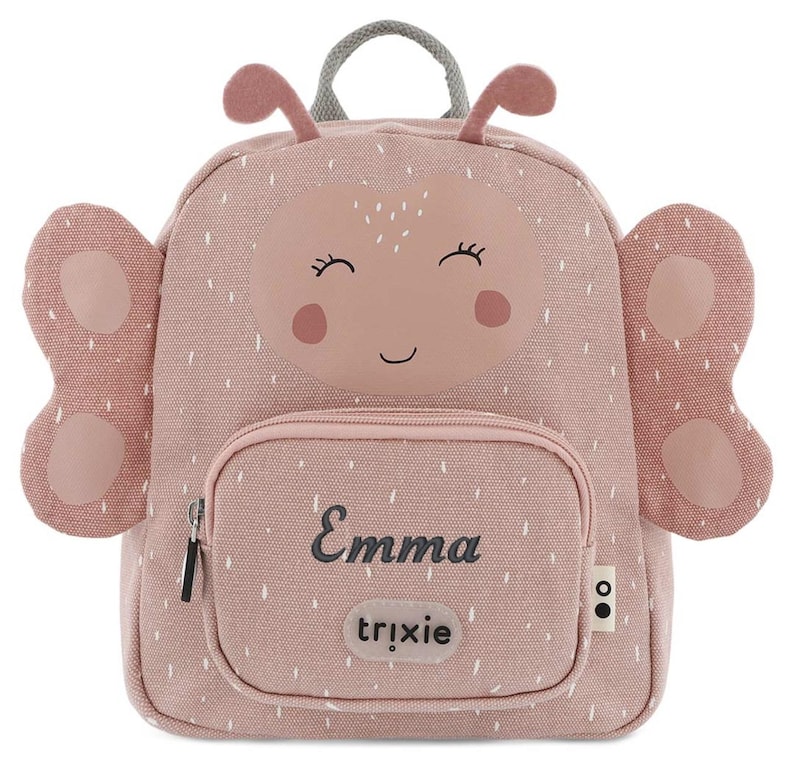 May include: A pink backpack with a butterfly design. The backpack features a smiling face, antennae, and butterfly wings. The name "Emma" is printed on the front pocket, with the brand name "trixie" below it. The backpack has a top handle and a zipper closure.