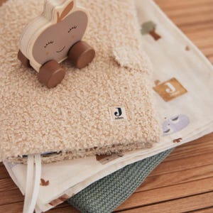 May include: A beige plush blanket with a wooden apple toy on top. The blanket has a white label with the text "Jollein" on it. The apple toy has a smiling face and wheels. The blanket is folded on top of a white and brown patterned blanket and a green blanket.