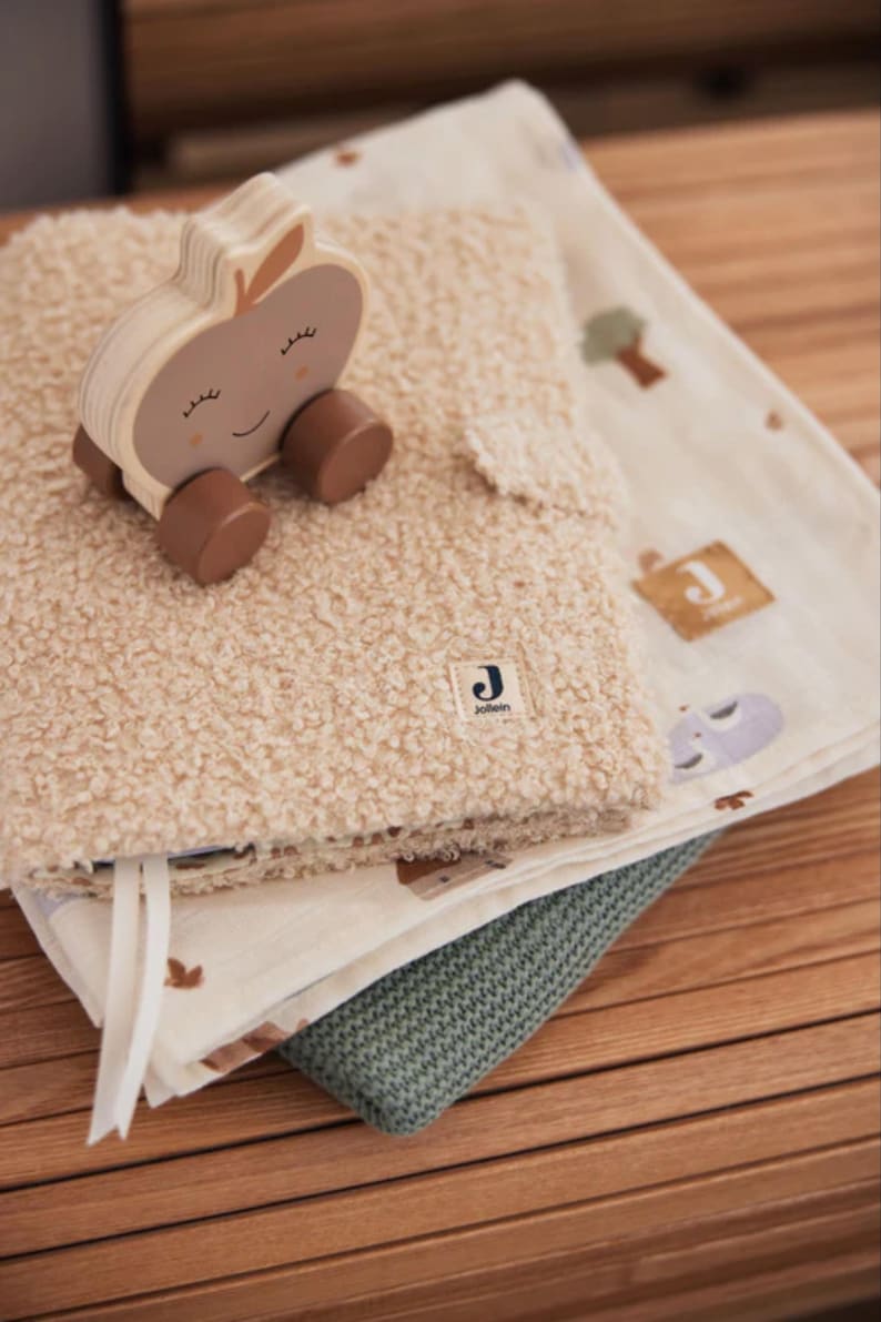 May include: A beige plush blanket with a wooden apple toy on top. The blanket has a white label with the text "Jollein" on it. The apple toy has a smiling face and wheels. The blanket is folded on top of a white and brown patterned blanket and a green blanket.