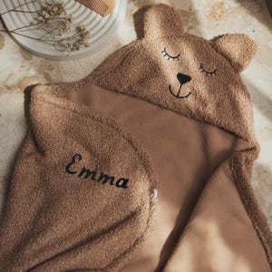 May include: A brown hooded baby towel with a bear face design. The towel features a hood with embroidered eyes, nose, and mouth. The name "Emma" is embroidered in black. A wooden comb and decorative sprigs are in the background.