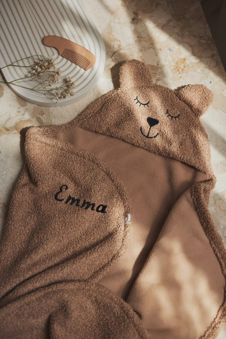 May include: A brown hooded baby towel with a bear face design. The towel features a hood with embroidered eyes, nose, and mouth. The name "Emma" is embroidered in black. A wooden comb and decorative sprigs are in the background.
