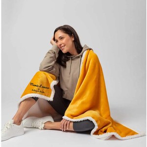 Puede incluir: Una manta de forro polar amarilla con ribete de sherpa blanco. La manta tiene el texto "Mamie d'amour" bordado en negro.