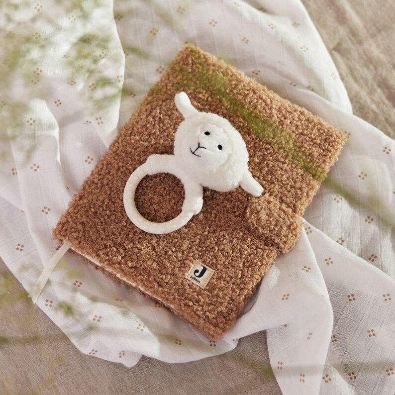 May include: A brown fuzzy book with a white plush lamb toy attached to the cover. The lamb has a ring attached to its body. The book has a letter "J" on the cover.