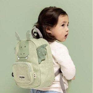 May include: A light green backpack with a dragon design, featuring a smiling face, wings, and small white dots. The backpack has a small front pocket and adjustable straps. The brand name "trixie" is visible on the front pocket.