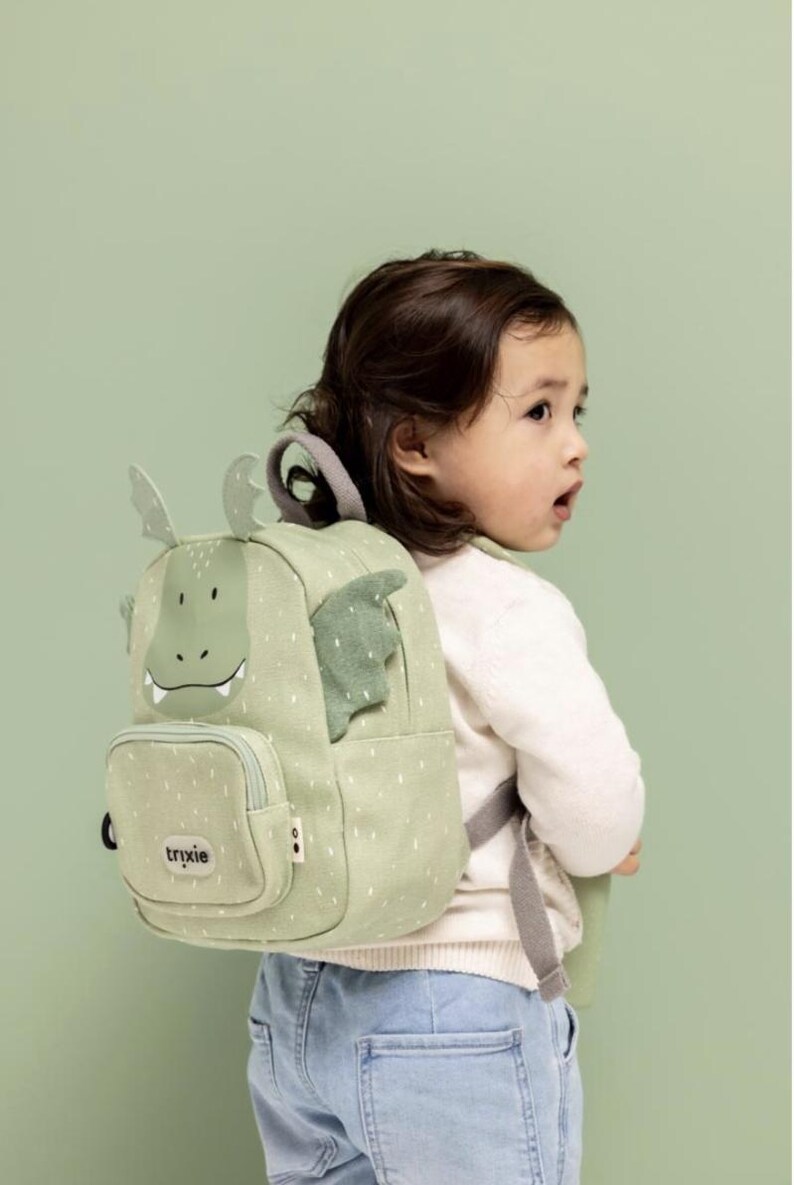 May include: A light green backpack with a dragon design, featuring a smiling face, wings, and small white dots. The backpack has a small front pocket and adjustable straps. The brand name "trixie" is visible on the front pocket.
