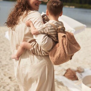 May include: A woman wearing a white baseball cap and a beige dress is carrying a child wearing a striped shirt and a brown fuzzy backpack. The child is looking at the woman and smiling.