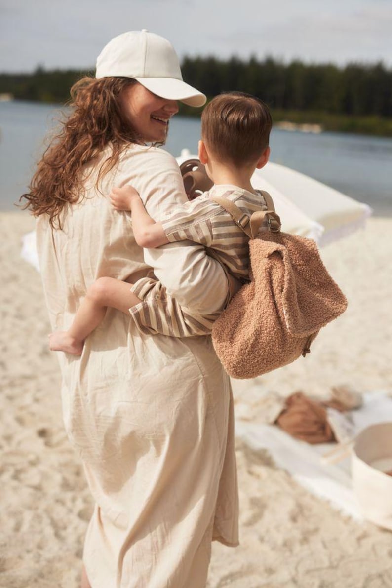 May include: A woman wearing a white baseball cap and a beige dress is carrying a child wearing a striped shirt and a brown fuzzy backpack. The child is looking at the woman and smiling.