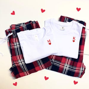 Puede incluir: Conjunto de pijama a juego con camisetas blancas con bordado de corazón rojo e iniciales, combinado con pantalones de pijama a cuadros rojos, blancos y azul marino. Acentos decorativos de corazón rojo dispersos.