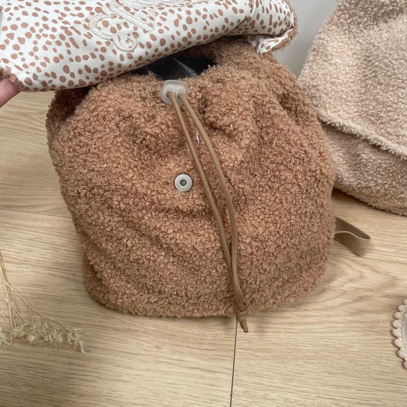 May include: A brown faux fur backpack with a drawstring closure and a white button. The backpack has a white and brown patterned lining.