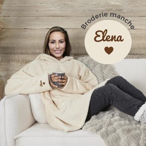May include: A cream-colored hooded blanket with the word "Elena" embroidered on the sleeve and a heart. The blanket is worn by a person sitting on a couch.
