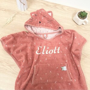 May include: A coral pink hooded towel with a cat face on the hood. The towel has a pocket on the front and the name "Eliott" embroidered on the front.