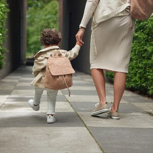 May include: A young child wearing a brown jacket and a fuzzy brown backpack walks with an adult. The child is holding the adult's hand. They are walking on a paved path with green hedges on either side.