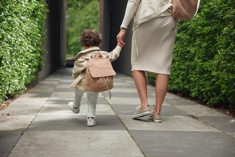 May include: A young child wearing a brown jacket and a fuzzy brown backpack walks with an adult. The child is holding the adult's hand. They are walking on a paved path with green hedges on either side.