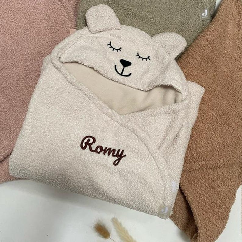May include: A light beige baby hooded towel with a bear face design on the hood. The towel has the name "Romy" embroidered in brown script. The towel is surrounded by other soft, textured fabrics in shades of pink, green, and brown.