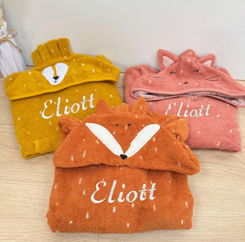 May include: Three hooded towels with animal faces. The towels are yellow, orange, and pink. Each towel has the name "Eliott" embroidered on it.