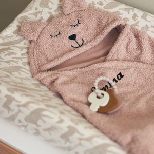 May include: A pink baby hooded towel with bear ears and embroidered eyes and nose. The towel is on a changing mat with a neutral animal print. A wooden and white teething ring is attached to the towel.