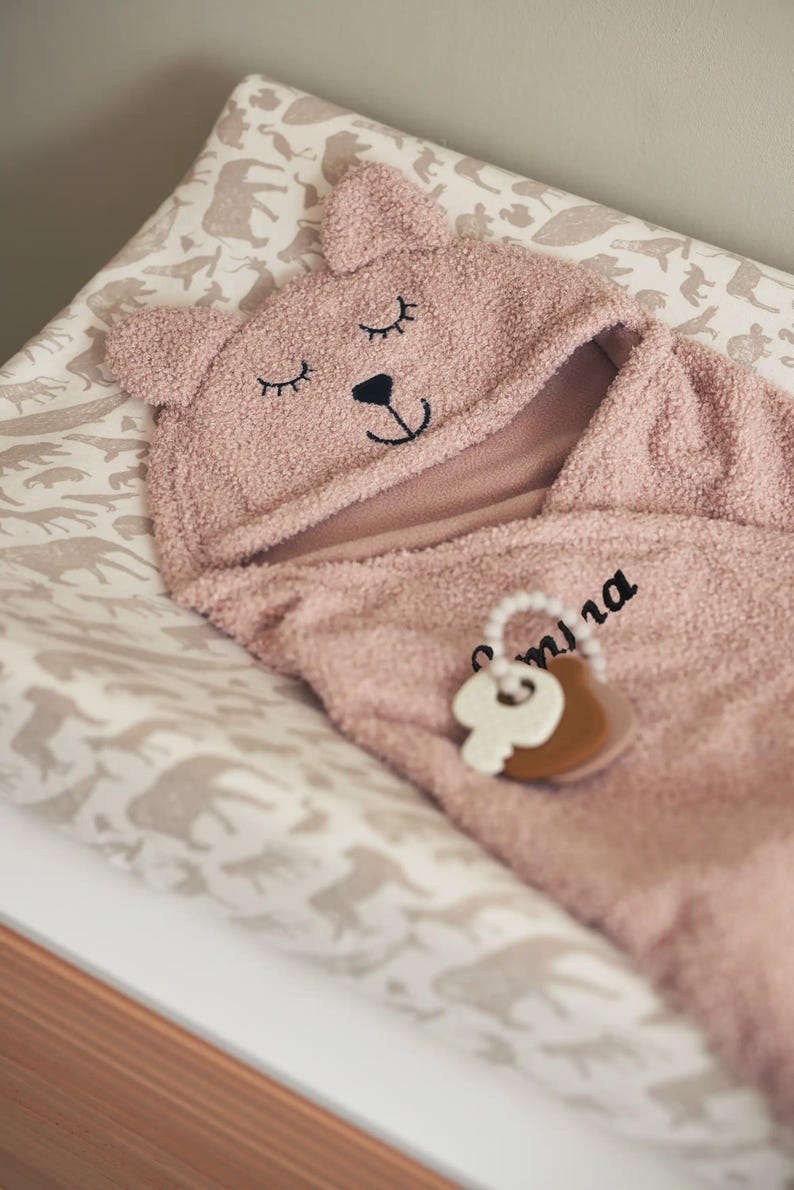 May include: A pink baby hooded towel with bear ears and embroidered eyes and nose. The towel is on a changing mat with a neutral animal print. A wooden and white teething ring is attached to the towel.