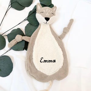 May include: A beige knitted cat-shaped security blanket with a white plush belly. The blanket has a knotted tail and embroidered eyes and nose. The name "Emma" is embroidered on the belly.