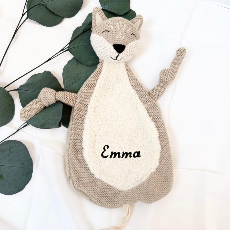 May include: A beige knitted cat-shaped security blanket with a white plush belly. The blanket has a knotted tail and embroidered eyes and nose. The name "Emma" is embroidered on the belly.