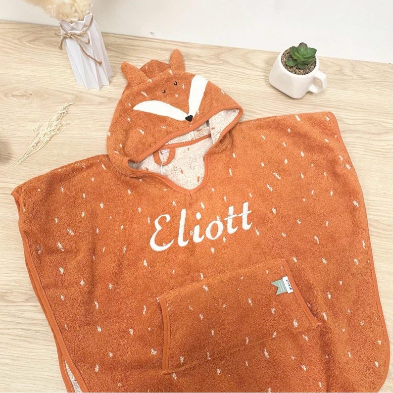 May include: Orange hooded towel with a fox face on the hood. The towel has a pocket and is embroidered with the name "Eliott".