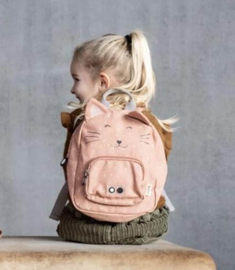 May include: A pink backpack with a cat face design. The backpack has a front pocket, a top handle, and adjustable straps. The cat face features whiskers, eyes, and ears. The backpack is worn by a child.