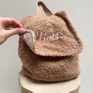 May include: A brown sherpa backpack with a white and brown polka dot lining. The backpack has a flap closure and the name "Elliott" is embroidered on the front.
