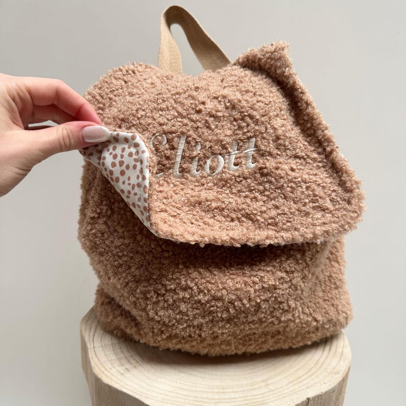 May include: A brown sherpa backpack with a white and brown polka dot lining. The backpack has a flap closure and the name "Elliott" is embroidered on the front.