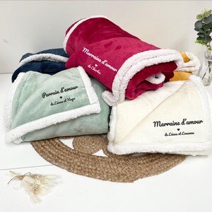 Puede incluir: Varias mantas suaves en tonos como el verde azulado, el burdeos y el crema. Cada manta tiene un ribete de sherpa blanco y texto bordado en negro. Están dispuestas sobre una estera tejida, con un pequeño arreglo floral en el fondo.