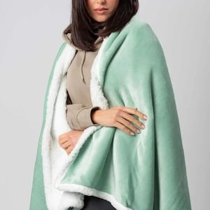May include: A soft, plush throw blanket with a light green outer layer and a white sherpa lining. The blanket is draped over a person's shoulders.