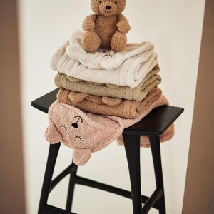 May include: A pile of soft baby towels in shades of pink, beige, and green, with bear-shaped hoods, and a small brown teddy bear on top. The towels are stacked on a black stool.