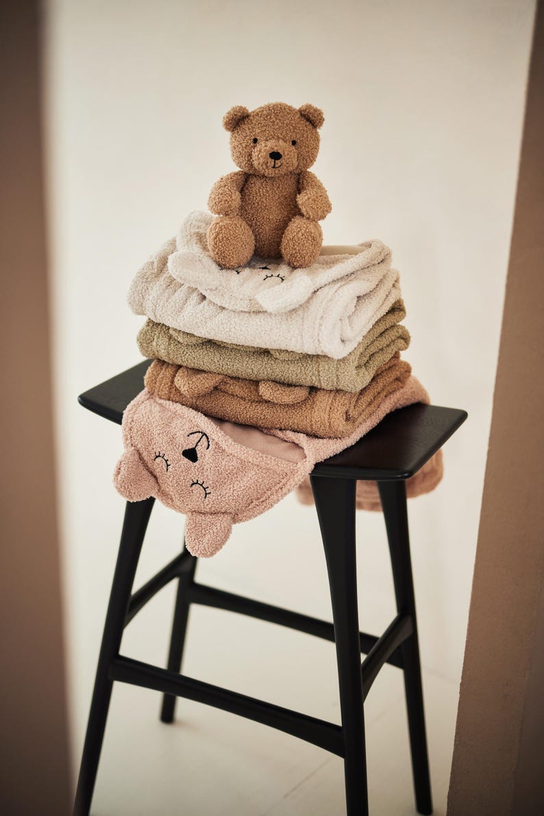 May include: A pile of soft baby towels in shades of pink, beige, and green, with bear-shaped hoods, and a small brown teddy bear on top. The towels are stacked on a black stool.