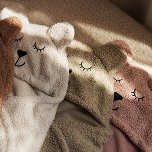 May include: Four teddy bear-shaped hooded towels in shades of brown, cream, and pink are arranged together. Each towel features embroidered closed eyes and a black nose, creating a cosy and whimsical design. The towels are made of soft, textured material.