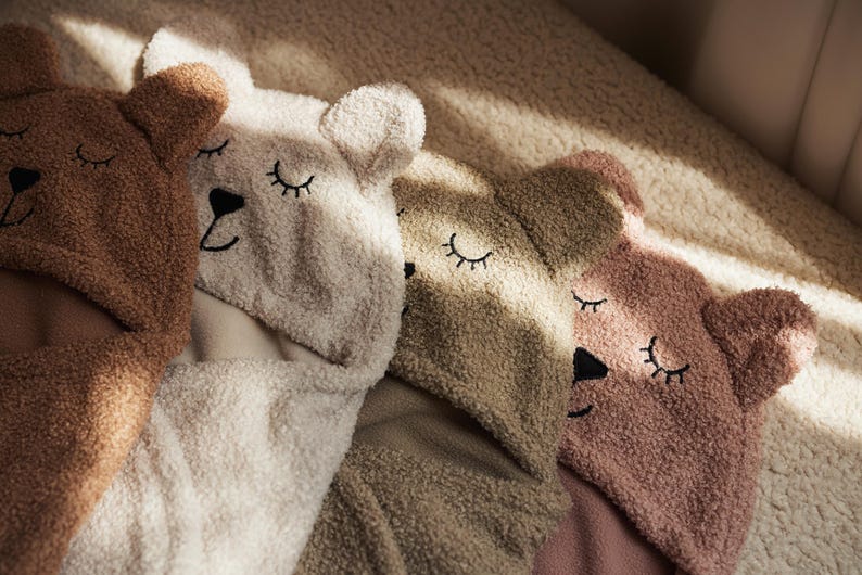 May include: Four teddy bear-shaped hooded towels in shades of brown, cream, and pink are arranged together. Each towel features embroidered closed eyes and a black nose, creating a cosy and whimsical design. The towels are made of soft, textured material.