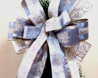 Puede incluir: Un lazo decorativo para árbol de Navidad. El lazo presenta capas de cintas plateadas y azul claro con adornos dorados y diseños de copos de nieve. El lazo está sujeto a una pequeña sección de un árbol de Navidad verde.