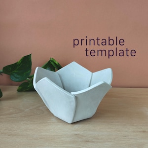 Puede incluir: Un cuenco de color gris claro de cinco lados con forma de flor y bordes acampanados. El texto "printable template" aparece en letras de color morado oscuro sobre el cuenco. El objeto tiene un acabado liso y mate y una abertura amplia.