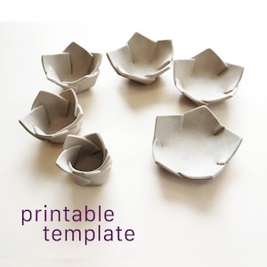 Può includere: Sei ciotole grigio chiaro a forma di fiore di varie dimensioni disposte in cerchio. Ogni ciotola presenta bordi piegati simili a petali e una finitura liscia e opaca. Il testo "printable template" è stampato in viola in basso a sinistra.