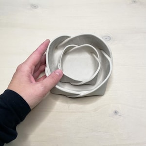 May include: A set of three nested, light gray ceramic bowls. The bowls are in a unique, intertwined design, with a smooth, matte finish. The bowls are of varying sizes, with the smallest bowl fitting inside the medium bowl, and the medium bowl fitting inside the largest bowl.
