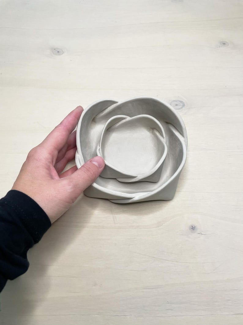 May include: A set of three nested, light gray ceramic bowls. The bowls are in a unique, intertwined design, with a smooth, matte finish. The bowls are of varying sizes, with the smallest bowl fitting inside the medium bowl, and the medium bowl fitting inside the largest bowl.